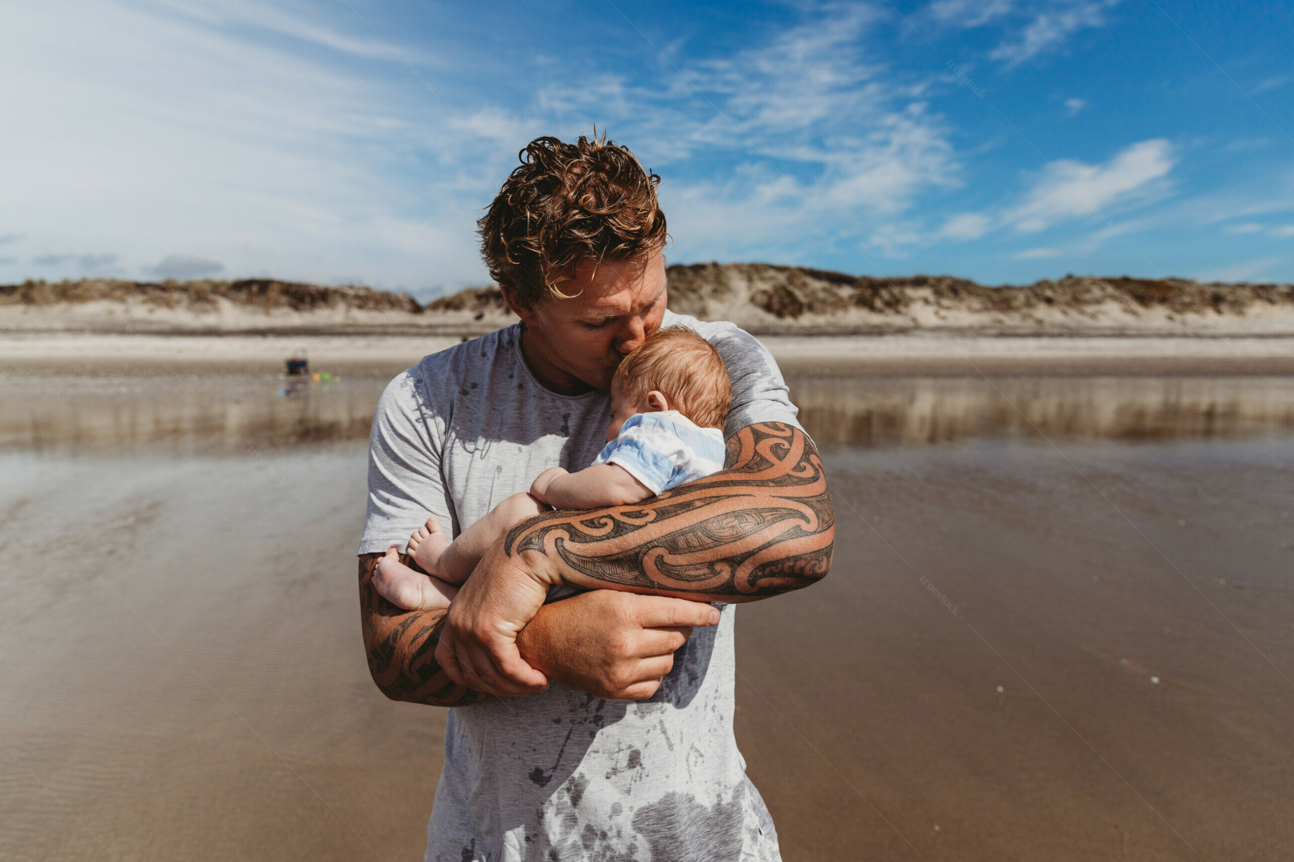 A stock image of a man with Māori tattoos, standing on a New Zealand beach holding a baby.